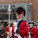 A group of historical reenactors dressed in 18th-century British or colonial military uniforms are gathered outside the Fort Pitt Museum. The central figure, smiling broadly, wears a red coat with blue facings, a tall bearskin cap, white gloves, and carries a halberd or spontoon. The scene appears to be a ceremonial or commemorative event at the museum.
