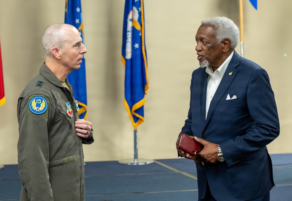 Two men are engaged in conversation at what appears to be a formal military ceremony or event. One man is wearing a military flight suit with patches including a '15th Air Force' insignia, while the other is dressed in a civilian navy blue suit. U.S. Air Force flags are visible in the background.