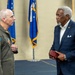 Two men are engaged in conversation at what appears to be a formal military ceremony or event. One man is wearing a military flight suit with patches including a '15th Air Force' insignia, while the other is dressed in a civilian navy blue suit. U.S. Air Force flags are visible in the background.