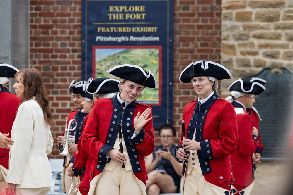 A group of historical reenactors dressed in 18th-century British or colonial military uniforms are performing or parading at Fort Pitt in Pittsburgh. They wear red coats with navy trim, tricorn hats, white wigs, and carry period instruments including flutes and a trumpet. The event appears to be tied to a featured exhibit called 'Pittsburgh's Revolution' visible on a sign behind them.