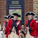 A group of historical reenactors dressed in 18th-century British or colonial military uniforms are performing or parading at Fort Pitt in Pittsburgh. They wear red coats with navy trim, tricorn hats, white wigs, and carry period instruments including flutes and a trumpet. The event appears to be tied to a featured exhibit called 'Pittsburgh's Revolution' visible on a sign behind them.