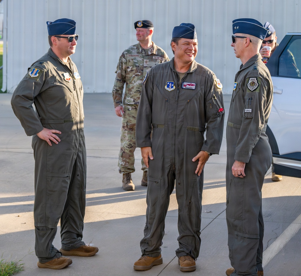 Three U.S. Air Force personnel in olive drab flight suits are engaged in conversation outdoors near what appears to be a hangar or flight line facility. The officer on the right displays two stars on his service cap, indicating the rank of Major General. A fourth individual in Army Combat Uniform (ACU) is visible in the background.