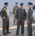 Three U.S. Air Force personnel in olive drab flight suits are engaged in conversation outdoors near what appears to be a hangar or flight line facility. The officer on the right displays two stars on his service cap, indicating the rank of Major General. A fourth individual in Army Combat Uniform (ACU) is visible in the background.