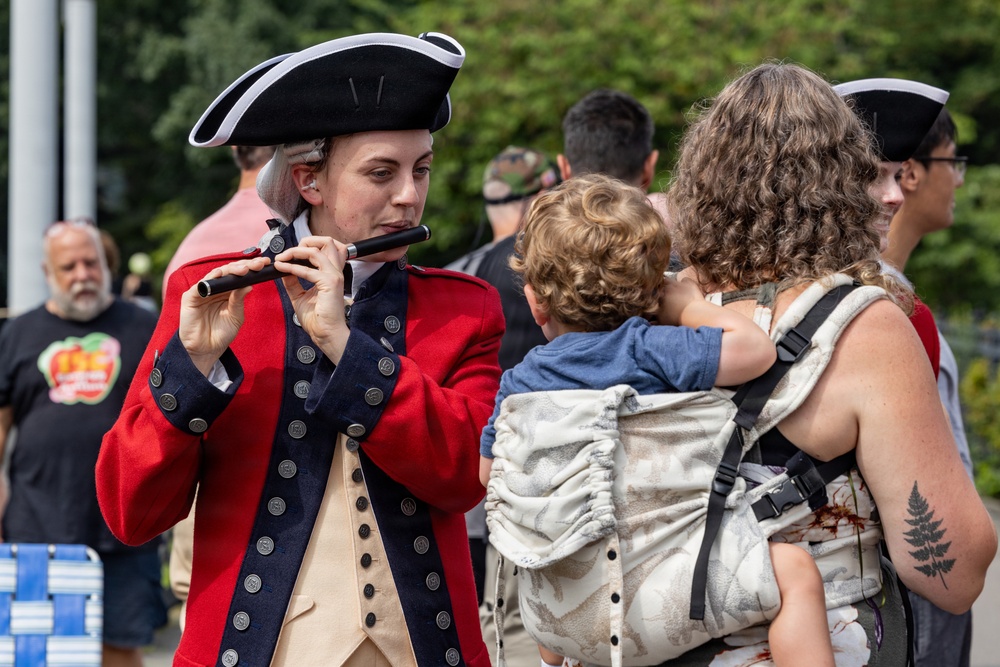 A historical reenactor dressed in 18th-century American Revolutionary War-era military uniform plays a fife at what appears to be an outdoor public event. A woman carrying a young child in a baby carrier stands nearby, seemingly watching or listening. The scene suggests a patriotic or historical festival setting with multiple attendees visible in the background.