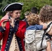 A historical reenactor dressed in 18th-century American Revolutionary War-era military uniform plays a fife at what appears to be an outdoor public event. A woman carrying a young child in a baby carrier stands nearby, seemingly watching or listening. The scene suggests a patriotic or historical festival setting with multiple attendees visible in the background.