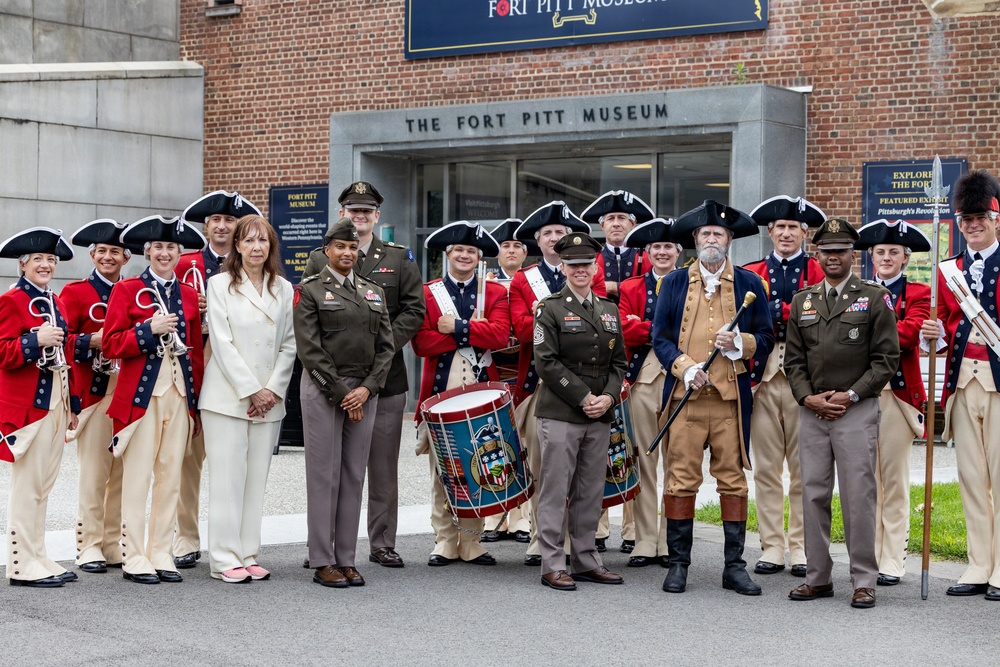 A group photo taken in front of The Fort Pitt Museum, featuring a mix of modern U.S. Army personnel in olive drab uniforms and historical reenactors dressed in 18th-century Revolutionary War-era colonial military attire. The group includes fife and drum corps members in red coats and tricorn hats, soldiers in contemporary Army service uniforms, and at least one civilian. The setting appears to be an official or ceremonial visit to the museum in Pittsburgh.