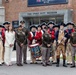 A group photo taken in front of The Fort Pitt Museum, featuring a mix of modern U.S. Army personnel in olive drab uniforms and historical reenactors dressed in 18th-century Revolutionary War-era colonial military attire. The group includes fife and drum corps members in red coats and tricorn hats, soldiers in contemporary Army service uniforms, and at least one civilian. The setting appears to be an official or ceremonial visit to the museum in Pittsburgh.