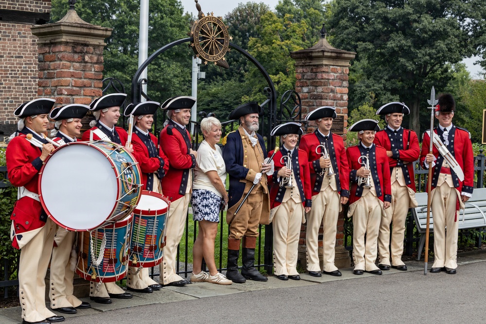 A group of performers dressed in 18th-century American Revolutionary War-era military uniforms pose for a photo at what appears to be a historic site. The group includes fife and drum corps members in red coats with tricorn hats, a colonial-era reenactor in tan/brown period clothing, and two civilians in modern dress. An ornamental iron gate with a circular emblem reading 'Daughters of the American Revolution' is visible in the background.