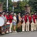 A group of performers dressed in 18th-century American Revolutionary War-era military uniforms pose for a photo at what appears to be a historic site. The group includes fife and drum corps members in red coats with tricorn hats, a colonial-era reenactor in tan/brown period clothing, and two civilians in modern dress. An ornamental iron gate with a circular emblem reading 'Daughters of the American Revolution' is visible in the background.