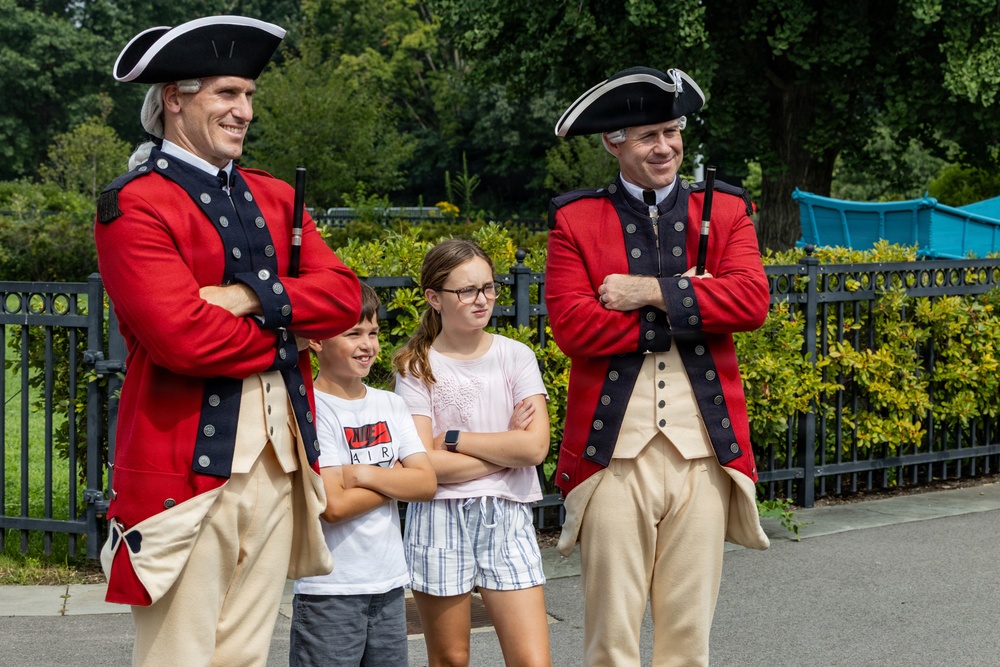 Two adults dressed in 18th-century British Redcoat military costumes pose with two children in what appears to be a theme park or historical attraction setting. The costumed performers wear red and navy coats with gold buttons, tricorn hats, and cream-colored breeches. The children are mimicking the crossed-arms pose of the performers, suggesting an interactive photo opportunity.