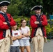 Two adults dressed in 18th-century British Redcoat military costumes pose with two children in what appears to be a theme park or historical attraction setting. The costumed performers wear red and navy coats with gold buttons, tricorn hats, and cream-colored breeches. The children are mimicking the crossed-arms pose of the performers, suggesting an interactive photo opportunity.
