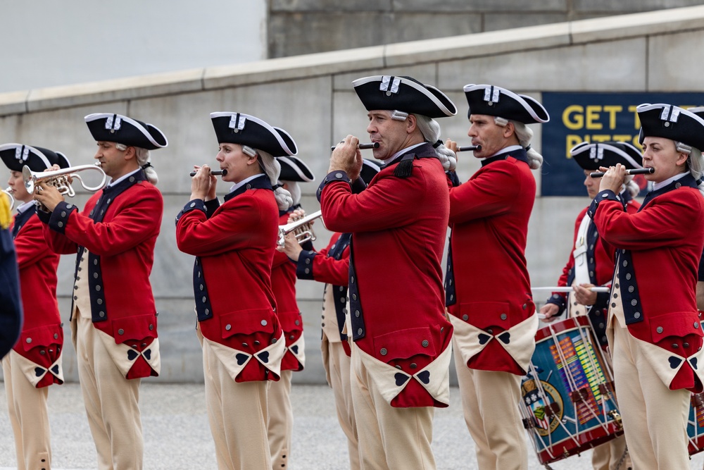 A group of performers dressed in 18th-century American Revolutionary War-era military uniforms are playing instruments in a marching band formation. They wear distinctive red coats, tricorn hats, white wigs, and cream-colored breeches with black decorative accents. A partial banner in the background reads 'GET T... PITTS...' suggesting a Pittsburgh-area venue or event.