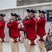 A group of performers dressed in 18th-century American Revolutionary War-era military uniforms are playing instruments in a marching band formation. They wear distinctive red coats, tricorn hats, white wigs, and cream-colored breeches with black decorative accents. A partial banner in the background reads 'GET T... PITTS...' suggesting a Pittsburgh-area venue or event.