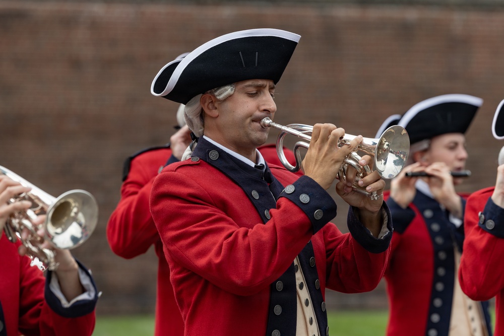 A group of musicians dressed in 18th-century colonial-era military uniforms are performing in what appears to be a historical reenactment or ceremonial event. The central figure is playing a silver bugle or trumpet while wearing a red coat, tricorn hat, and powdered wig. Additional musicians are visible in the background playing similar brass and wind instruments.