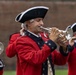 A group of musicians dressed in 18th-century colonial-era military uniforms are performing in what appears to be a historical reenactment or ceremonial event. The central figure is playing a silver bugle or trumpet while wearing a red coat, tricorn hat, and powdered wig. Additional musicians are visible in the background playing similar brass and wind instruments.