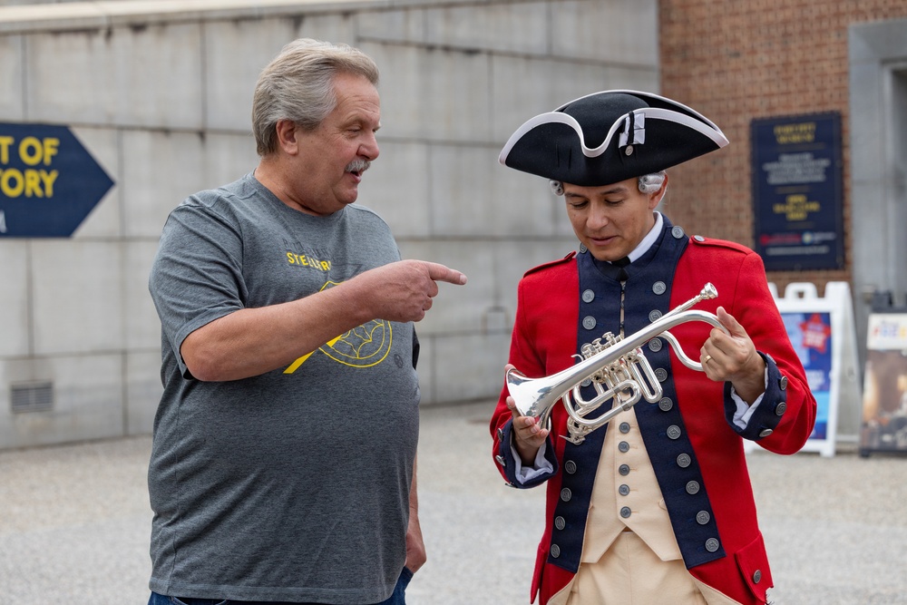 Two men are interacting outdoors near what appears to be a historical museum or visitor center. One man is dressed in a colonial-era costume including a tricorn hat and red military-style coat, holding a silver trumpet or cornet. The other man, wearing a Pittsburgh Steelers t-shirt, is pointing at the instrument with apparent interest or commentary.