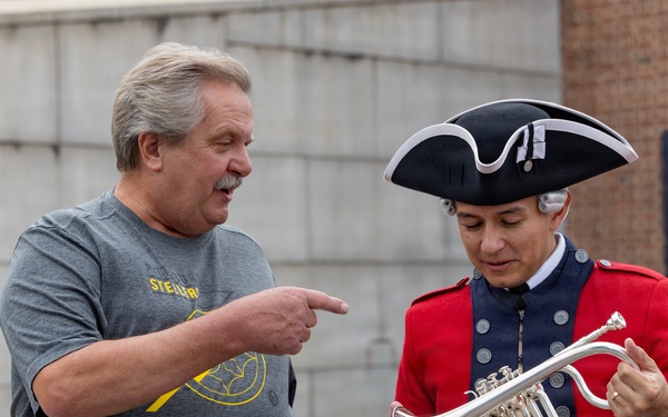 Two men are interacting outdoors near what appears to be a historical museum or visitor center. One man is dressed in a colonial-era costume including a tricorn hat and red military-style coat, holding a silver trumpet or cornet. The other man, wearing a Pittsburgh Steelers t-shirt, is pointing at the instrument with apparent interest or commentary.