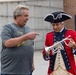 Two men are interacting outdoors near what appears to be a historical museum or visitor center. One man is dressed in a colonial-era costume including a tricorn hat and red military-style coat, holding a silver trumpet or cornet. The other man, wearing a Pittsburgh Steelers t-shirt, is pointing at the instrument with apparent interest or commentary.