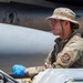 A U.S. Air Force service member in OCP (Operational Camouflage Pattern) uniform is seated at the controls of a ground support vehicle on a flight line. He is wearing a boonie hat, blue nitrile gloves, and orange ear protection, working in proximity to what appears to be a military jet aircraft. A unit patch is visible on his left sleeve, consistent with U.S. Air Forces markings.