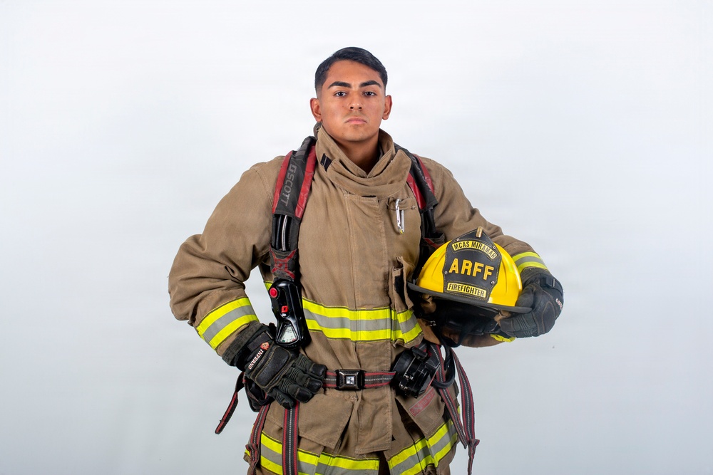 A young firefighter in full turnout gear poses against a white background in a professional portrait-style photograph. He is holding a yellow helmet labeled 'ARFF' and 'MCAS Miramar Firefighter', indicating he is an Aircraft Rescue and Firefighting specialist at Marine Corps Air Station Miramar. The image appears to be an official or promotional photograph.