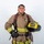 A young firefighter in full turnout gear poses against a white background in a professional portrait-style photograph. He is holding a yellow helmet labeled 'ARFF' and 'MCAS Miramar Firefighter', indicating he is an Aircraft Rescue and Firefighting specialist at Marine Corps Air Station Miramar. The image appears to be an official or promotional photograph.