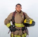 A young firefighter in full turnout gear poses against a white background in a professional portrait-style photograph. He is holding a yellow helmet labeled 'ARFF' and 'MCAS Miramar Firefighter', indicating he is an Aircraft Rescue and Firefighting specialist at Marine Corps Air Station Miramar. The image appears to be an official or promotional photograph.