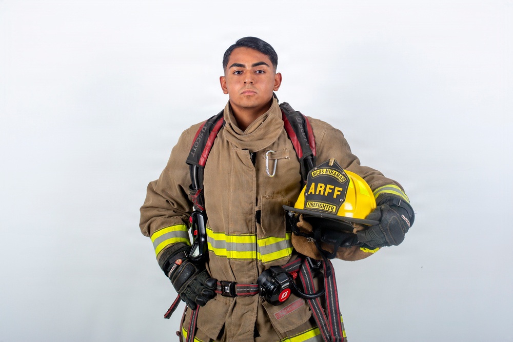 A firefighter in full turnout gear poses against a plain white background in what appears to be a professional portrait. The individual is holding a yellow helmet labeled 'ARFF Firefighter' and 'MCAS Miramar', indicating affiliation with Aircraft Rescue and Fire Fighting at Marine Corps Air Station Miramar. The subject is wearing complete structural firefighting gear including gloves, reflective striping, and SCBA harness straps.