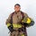 A firefighter in full turnout gear poses against a plain white background in what appears to be a professional portrait. The individual is holding a yellow helmet labeled 'ARFF Firefighter' and 'MCAS Miramar', indicating affiliation with Aircraft Rescue and Fire Fighting at Marine Corps Air Station Miramar. The subject is wearing complete structural firefighting gear including gloves, reflective striping, and SCBA harness straps.