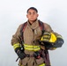 A firefighter in full turnout gear poses against a plain white background in what appears to be a professional portrait. The individual is holding a yellow helmet labeled 'ARFF Firefighter' and 'MCAS Miramar', indicating affiliation with Aircraft Rescue and Fire Fighting at Marine Corps Air Station Miramar. The subject is wearing complete structural firefighting gear including gloves, reflective striping, and SCBA harness straps.