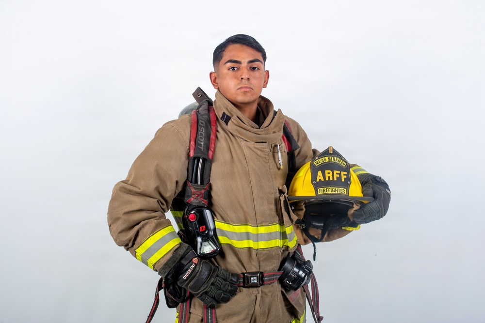 A young firefighter poses in full turnout gear against a white studio background. He is holding a yellow helmet and wearing a complete structural firefighting ensemble with SCBA equipment. The helmet and gear indicate affiliation with MCAS Miramar ARFF (Aircraft Rescue and Firefighting).