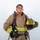 A young firefighter poses in full turnout gear against a white studio background. He is holding a yellow helmet and wearing a complete structural firefighting ensemble with SCBA equipment. The helmet and gear indicate affiliation with MCAS Miramar ARFF (Aircraft Rescue and Firefighting).