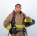 A young firefighter poses in full turnout gear against a white studio background. He is holding a yellow helmet and wearing a complete structural firefighting ensemble with SCBA equipment. The helmet and gear indicate affiliation with MCAS Miramar ARFF (Aircraft Rescue and Firefighting).