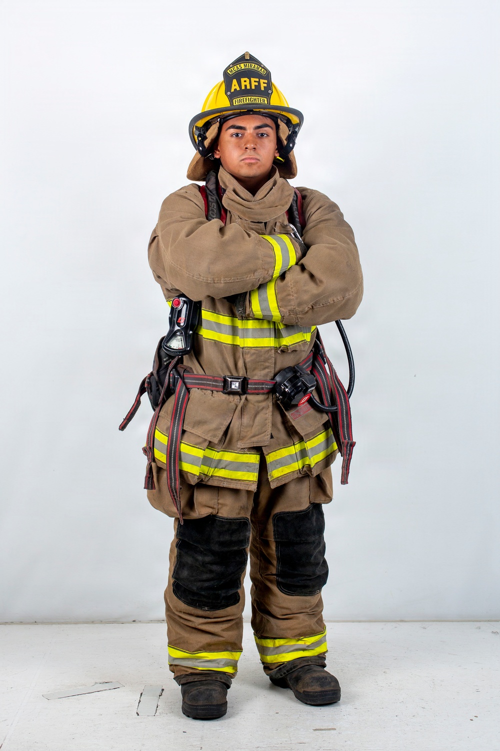 A firefighter in full turnout gear stands in a formal portrait pose with arms crossed against a white studio background. The individual is wearing an ARFF (Aircraft Rescue and Firefighting) helmet marked 'MCAS Miramar' and 'Firefighter,' indicating affiliation with Marine Corps Air Station Miramar. The gear is tan/brown with high-visibility yellow-green reflective striping and includes SCBA harness equipment.