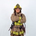 A firefighter in full turnout gear stands in a formal portrait pose with arms crossed against a white studio background. The individual is wearing an ARFF (Aircraft Rescue and Firefighting) helmet marked 'MCAS Miramar' and 'Firefighter,' indicating affiliation with Marine Corps Air Station Miramar. The gear is tan/brown with high-visibility yellow-green reflective striping and includes SCBA harness equipment.
