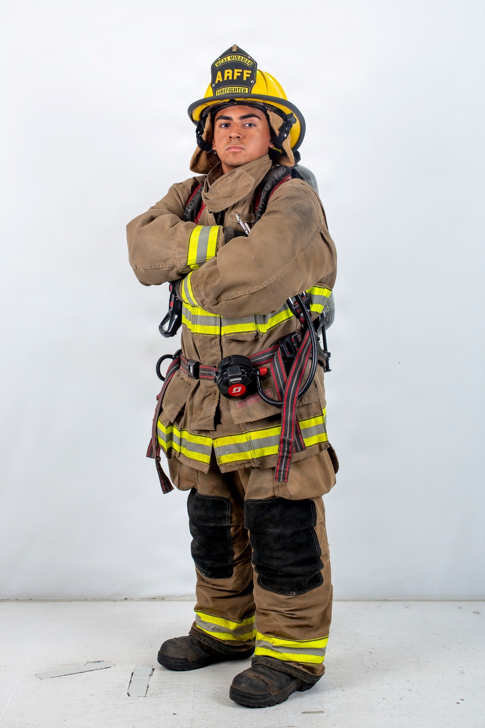 A firefighter in full turnout gear poses against a white background with arms crossed in a confident stance. The helmet displays 'ARFF FIREFIGHTER' and 'MCAS MIRAMAR', identifying this individual as an Aircraft Rescue and Firefighting specialist at Marine Corps Air Station Miramar. The subject is fully equipped with SCBA harness, reflective striping, and firefighting boots.