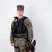 A U.S. Marine in MARPAT woodland digital camouflage uniform stands in a formal portrait pose with a military working dog, likely a Belgian Malinois. The Marine is wearing a tactical vest with a badge, body camera, and other law enforcement equipment, indicating a Military Police or K-9 handler role. The dog is seated beside the handler wearing a black tactical harness.