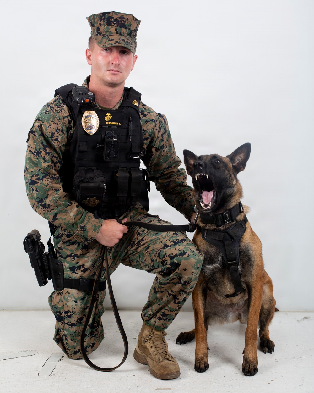A U.S. Marine Corps Military Police sergeant in MARPAT digital camouflage uniform kneels beside a Belgian Malinois military working dog. The Marine is wearing a tactical vest with a badge, name tape reading 'FESENMAIER M.', and is armed with a sidearm. The dog is wearing a tactical harness and leash and has its mouth open in an alert/barking posture.