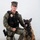 MCAS Marines pose for content 2026 A U.S. Marine Corps Military Police sergeant in MARPAT digital camouflage uniform kneels beside a Belgian Malinois military working dog. The Marine is wearing a tactical vest with a badge, name tape reading 'FESENMAIER M.', and is armed with a sidearm. The dog is wearing a tactical harness and leash and has its mouth open in an alert/barking posture.