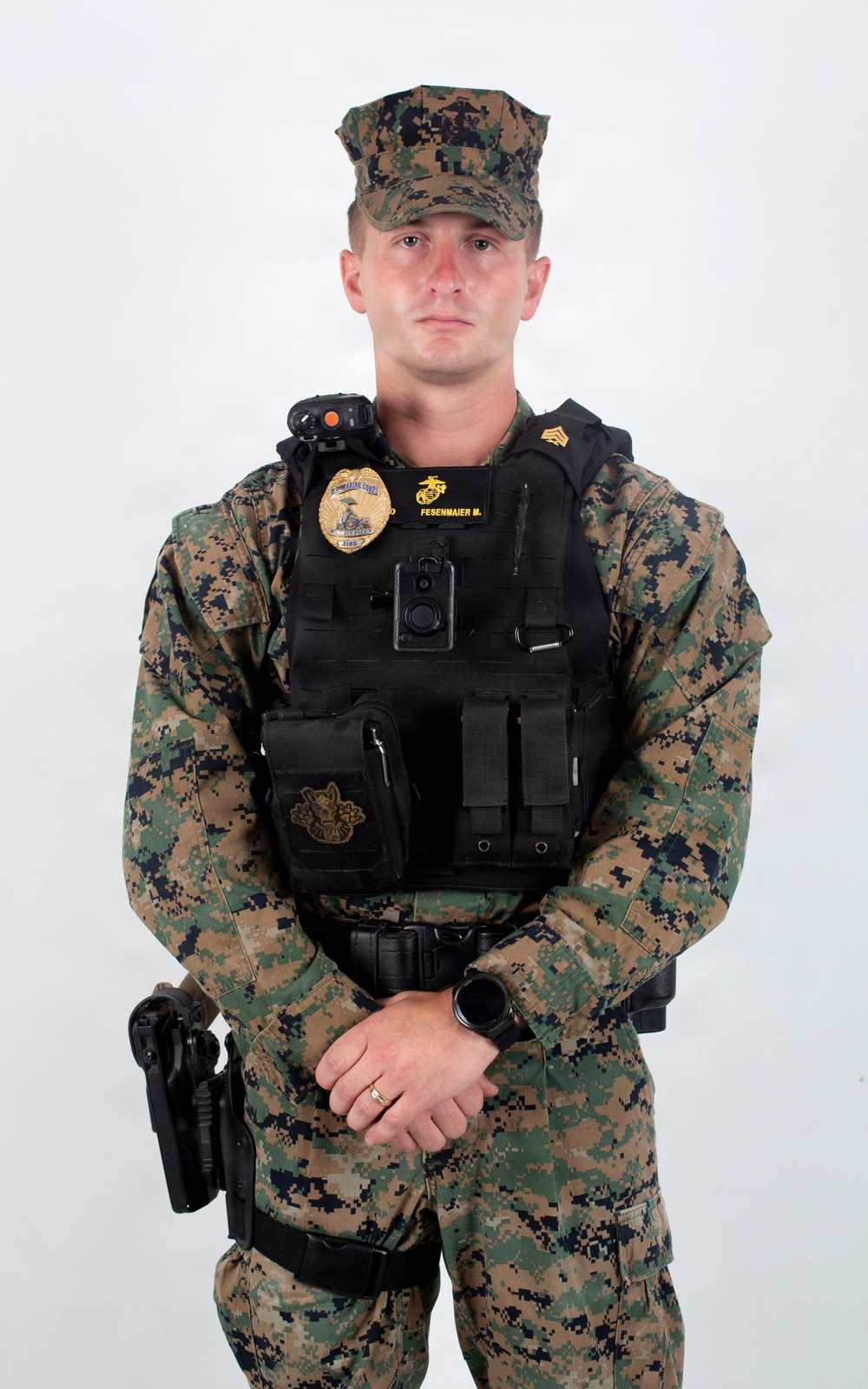 A U.S. Marine Corps Sergeant in woodland MARPAT digital camouflage uniform is posed for an official portrait against a white background. The individual is wearing a black tactical vest with a law enforcement badge, body camera, and name tape reading 'FESENMAIER M.' indicating a Military Police or provost marshal role. The subject stands in a formal at-ease position with hands clasped in front.