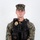 MCAS Marines pose for content 2026 A U.S. Marine Corps Sergeant in woodland MARPAT digital camouflage uniform is posed for an official portrait against a white background. The individual is wearing a black tactical vest with a law enforcement badge, body camera, and name tape reading 'FESENMAIER M.' indicating a Military Police or provost marshal role. The subject stands in a formal at-ease position with hands clasped in front.
