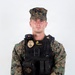 A U.S. Marine Corps Sergeant in woodland MARPAT digital camouflage uniform is posed for an official portrait against a white background. The individual is wearing a black tactical vest with a law enforcement badge, body camera, and name tape reading 'FESENMAIER M.' indicating a Military Police or provost marshal role. The subject stands in a formal at-ease position with hands clasped in front.