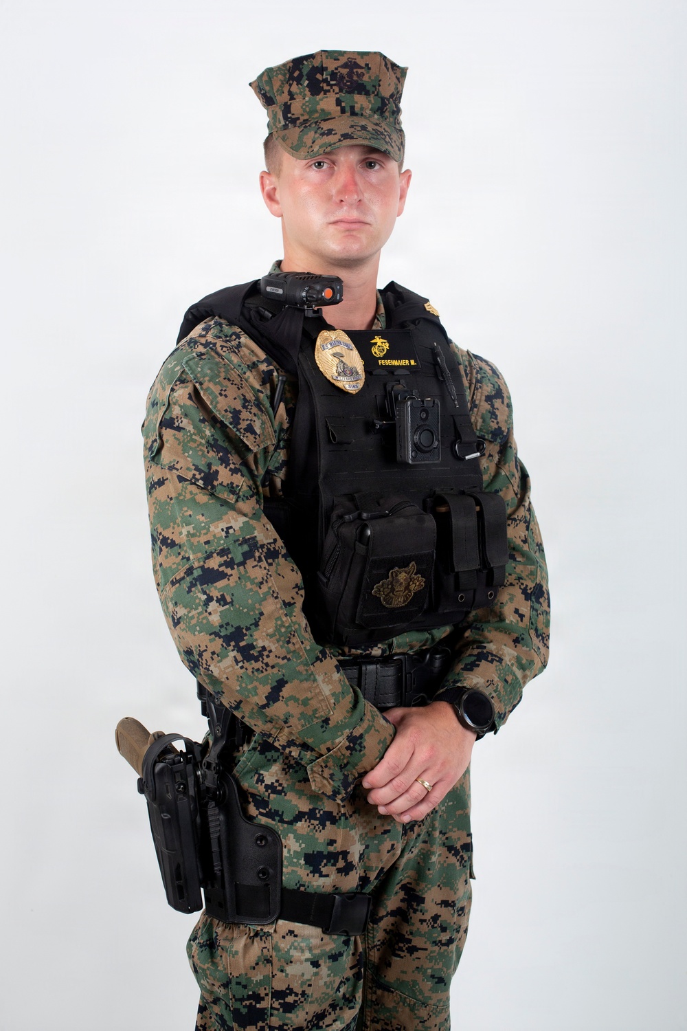 A male service member in U.S. Marine Corps MARPAT woodland digital camouflage uniform is posed against a white studio background. He is wearing a black tactical vest with a law enforcement badge, name tape reading 'FESENMAIER M.', a body camera, and an Eagle Globe and Anchor emblem. He is equipped with a sidearm in a holster on his left hip and appears to be a U.S. Marine Corps Military Police officer.