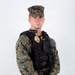 A male service member in U.S. Marine Corps MARPAT woodland digital camouflage uniform is posed against a white studio background. He is wearing a black tactical vest with a law enforcement badge, name tape reading 'FESENMAIER M.', a body camera, and an Eagle Globe and Anchor emblem. He is equipped with a sidearm in a holster on his left hip and appears to be a U.S. Marine Corps Military Police officer.