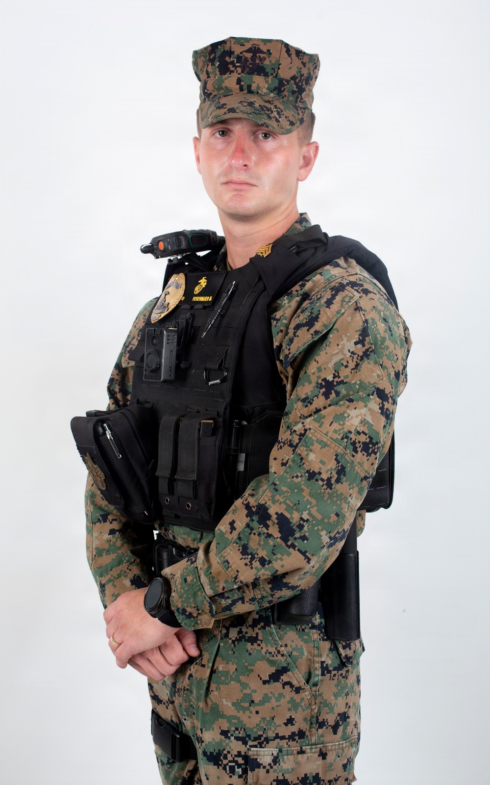 A male service member is posed against a plain white background in a formal portrait-style photograph. He is wearing USMC MARPAT woodland camouflage utilities with a black tactical vest/plate carrier over the top, along with various law enforcement or security equipment. The image appears to be an official or professional identification-style photograph.