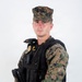 A male service member is posed against a plain white background in a formal portrait-style photograph. He is wearing USMC MARPAT woodland camouflage utilities with a black tactical vest/plate carrier over the top, along with various law enforcement or security equipment. The image appears to be an official or professional identification-style photograph.