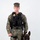 MCAS Marines pose for content 2026 A U.S. Marine in MARPAT woodland digital camouflage uniform stands at attention alongside a Belgian Malinois military working dog. The Marine is wearing a tactical plate carrier vest with a badge, name tape reading 'FESENMAYER', and a body camera, indicating a military police or provost marshal role. The dog is seated in a black tactical harness, suggesting this is an official K-9 unit portrait.