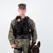 A U.S. Marine in MARPAT woodland digital camouflage uniform stands at attention alongside a Belgian Malinois military working dog. The Marine is wearing a tactical plate carrier vest with a badge, name tape reading 'FESENMAYER', and a body camera, indicating a military police or provost marshal role. The dog is seated in a black tactical harness, suggesting this is an official K-9 unit portrait.