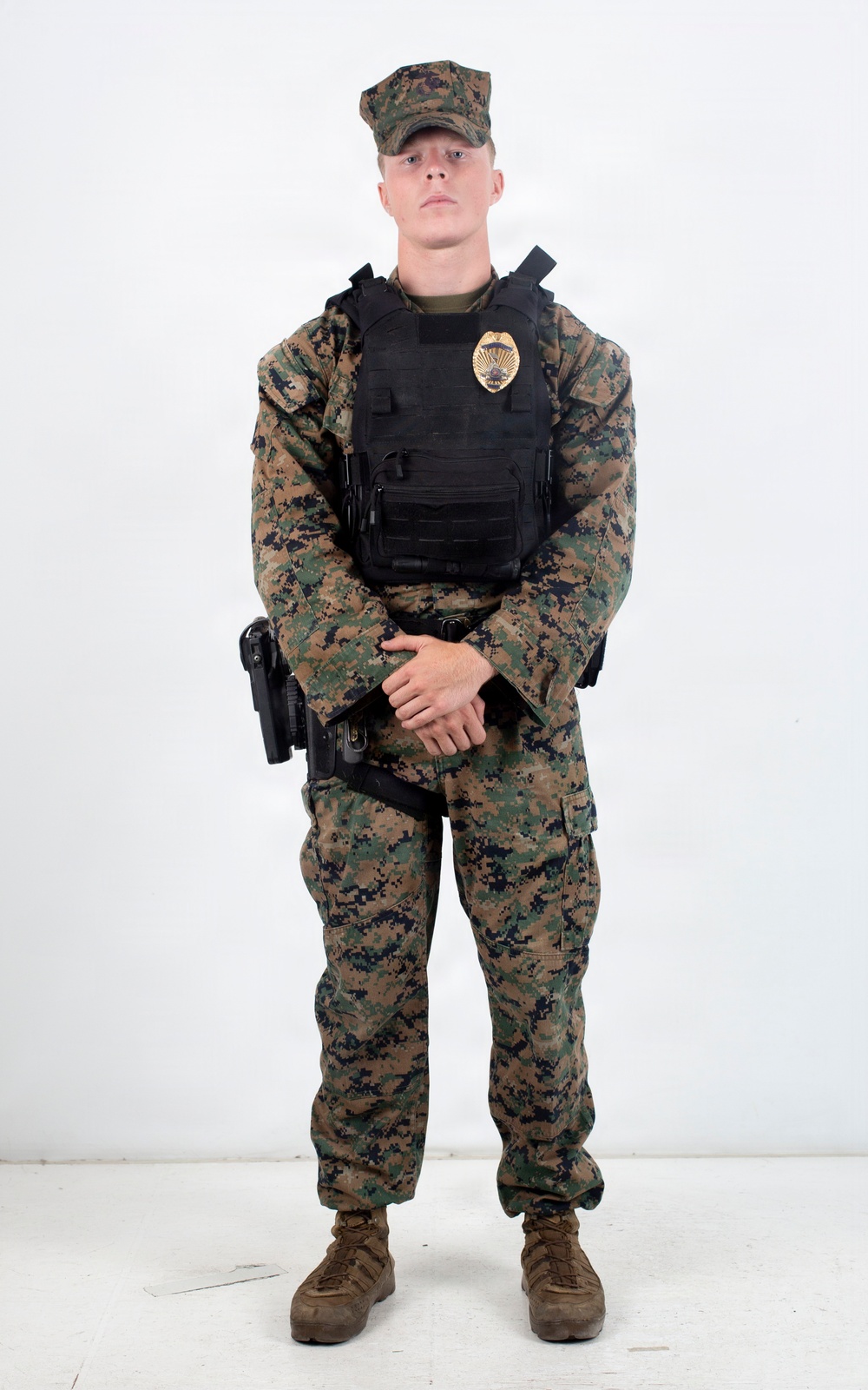 A young male in USMC MARPAT woodland digital camouflage uniform stands at ease against a plain white background. He is wearing a black tactical plate carrier vest with a gold badge affixed to the front, and a duty belt with a holstered sidearm. The image appears to be a formal or reference photograph, likely for law enforcement or military police documentation.