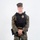 MCAS Marines pose for content 2026 A young male in USMC MARPAT woodland digital camouflage uniform stands at ease against a plain white background. He is wearing a black tactical plate carrier vest with a gold badge affixed to the front, and a duty belt with a holstered sidearm. The image appears to be a formal or reference photograph, likely for law enforcement or military police documentation.