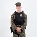 A young male in USMC MARPAT woodland digital camouflage uniform stands at ease against a plain white background. He is wearing a black tactical plate carrier vest with a gold badge affixed to the front, and a duty belt with a holstered sidearm. The image appears to be a formal or reference photograph, likely for law enforcement or military police documentation.